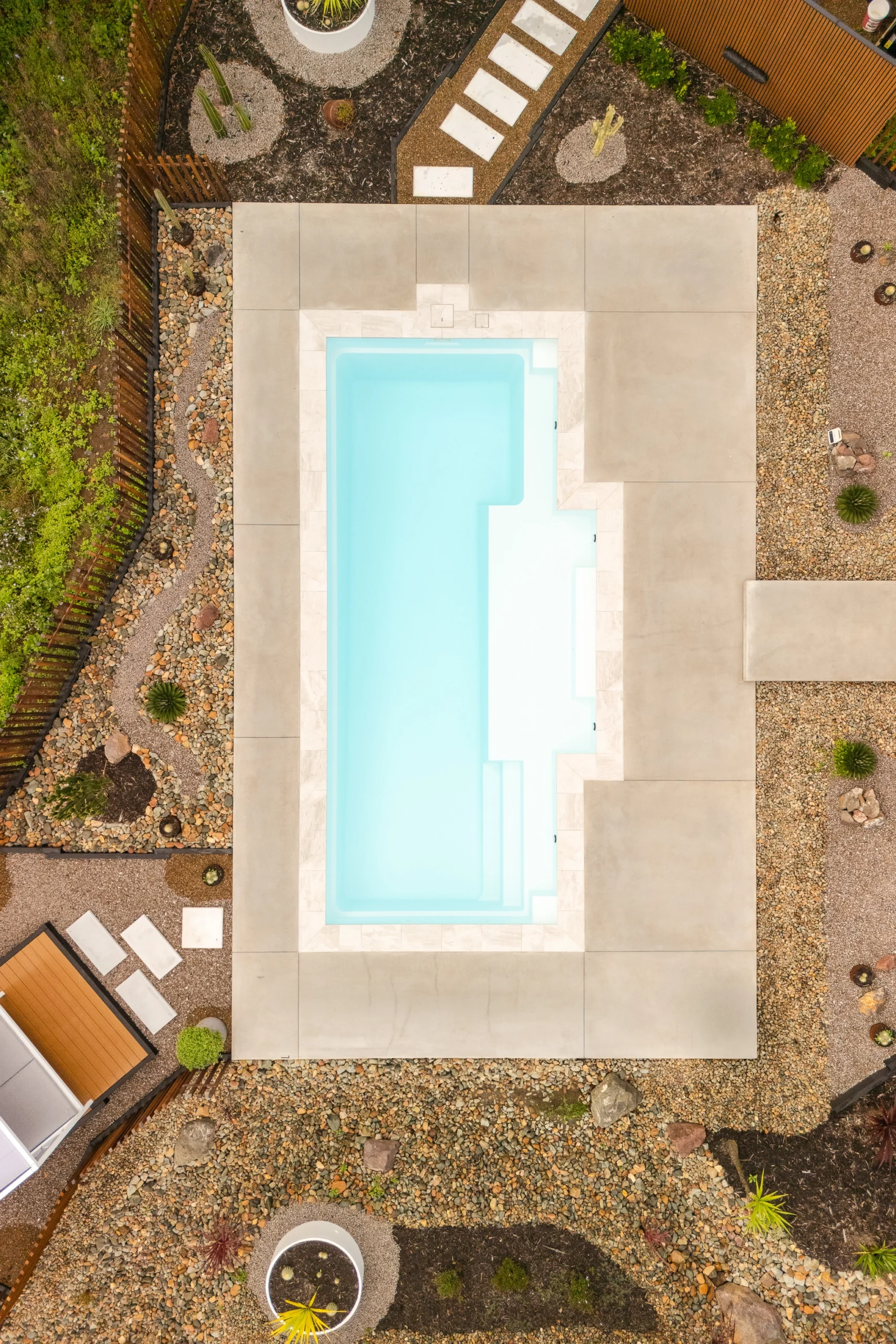 The pool installation of The Eternal 9.15m fibreglass swimming pool in Pearl is installed by Nexus Pools Sunshine Coast North. The pool is surrounded by light stone paving, decorative river pebbles and tropical landscaping, with timber fencing and lush greenery framing the backyard. The Pearl pool colour creates soft, pale blue water that complements the natural surroundings and tranquil waterway in the background. There is some cacti and a river in the background of the property.