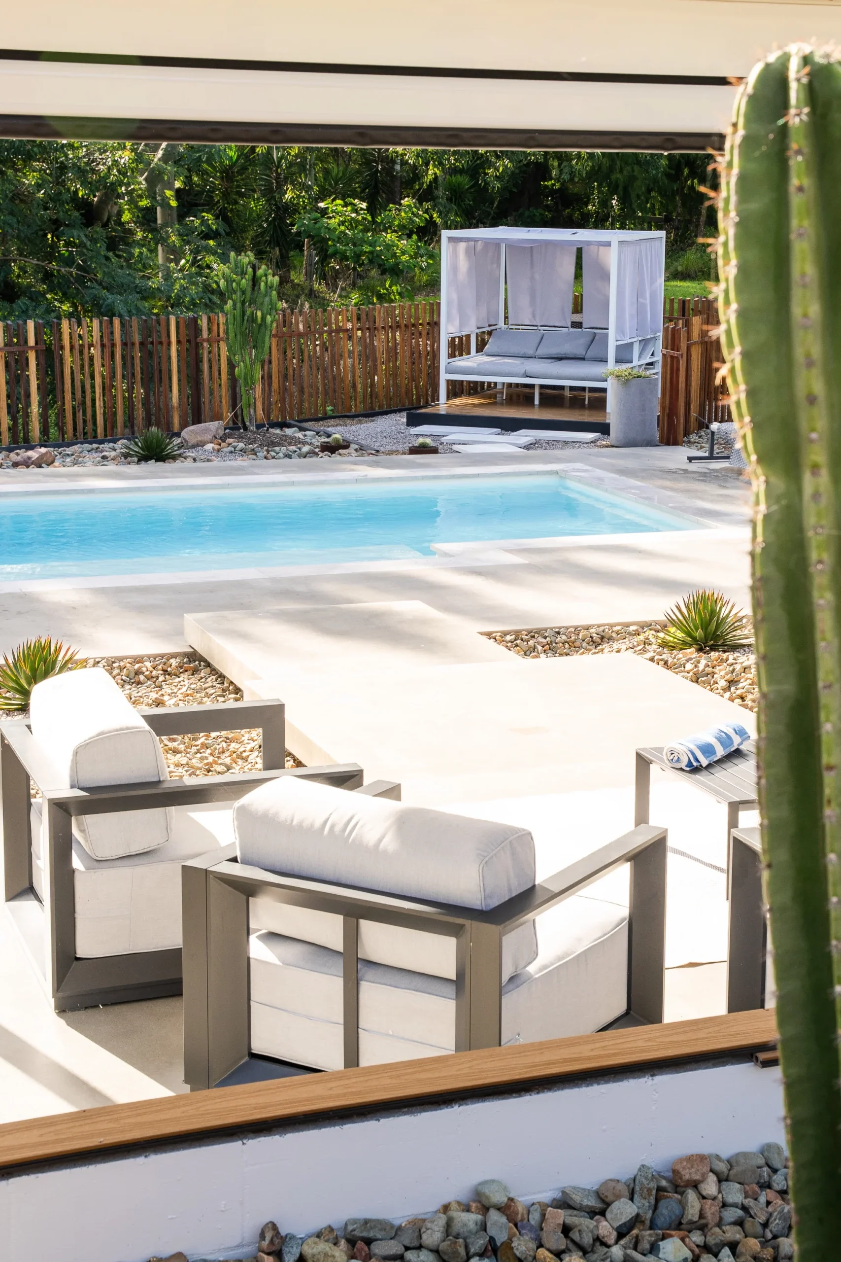 The Eternal 9.15m fibreglass swimming pool in Pearl is installed by Nexus Pools Sunshine Coast North. The pool is surrounded by light stone paving, decorative river pebbles and tropical landscaping, with timber fencing and lush greenery framing the backyard. The Pearl pool colour creates soft, pale blue water that complements the natural surroundings and tranquil waterway in the background. There is some cacti and a river in the background of the property.