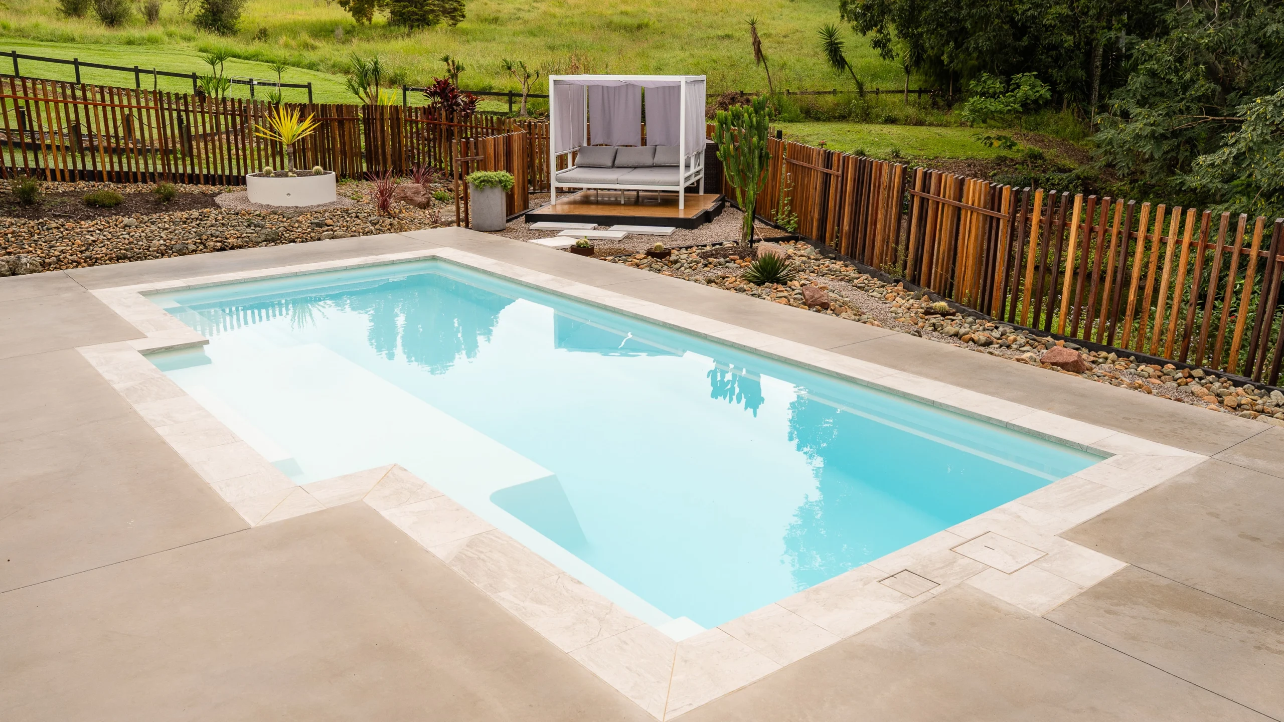 The pool installation of The Eternal 9.15m fibreglass swimming pool in Pearl is installed by Nexus Pools Sunshine Coast North. The pool is surrounded by light stone paving, decorative river pebbles and tropical landscaping, with timber fencing and lush greenery framing the backyard. The Pearl pool colour creates soft, pale blue water that complements the natural surroundings and tranquil waterway in the background. There is some cacti and a river in the background of the property.
